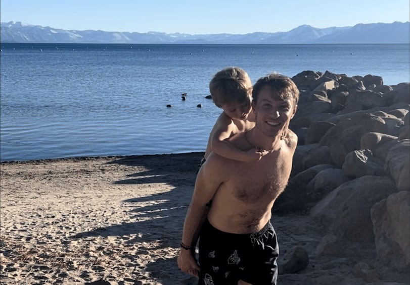 A person carrying a child on a rocky beach with a calm sea and mountains in the background. 2