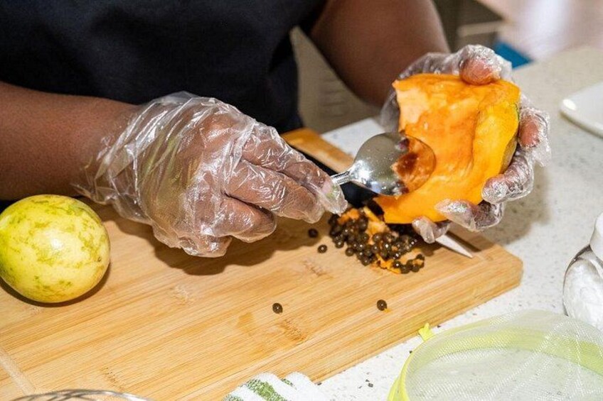 Une personne portant des gants manipule un fruit, peut-être une papaye, sur une planche à découper en bois. Une cuillère et quelques graines se trouvent à proximité. 3