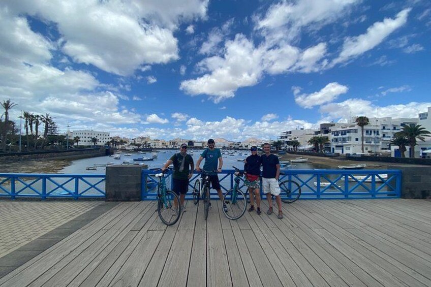 A wooden boardwalk with people and bicycles, overlooking a body of water with boats and buildings in the background. 3