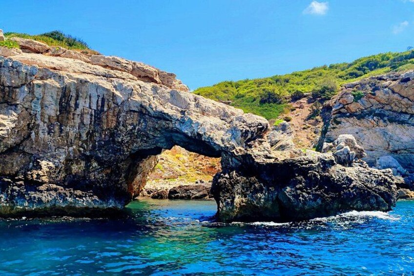 A natural rock formation with a cave-like opening, clear blue water, and green hills in the background. 2