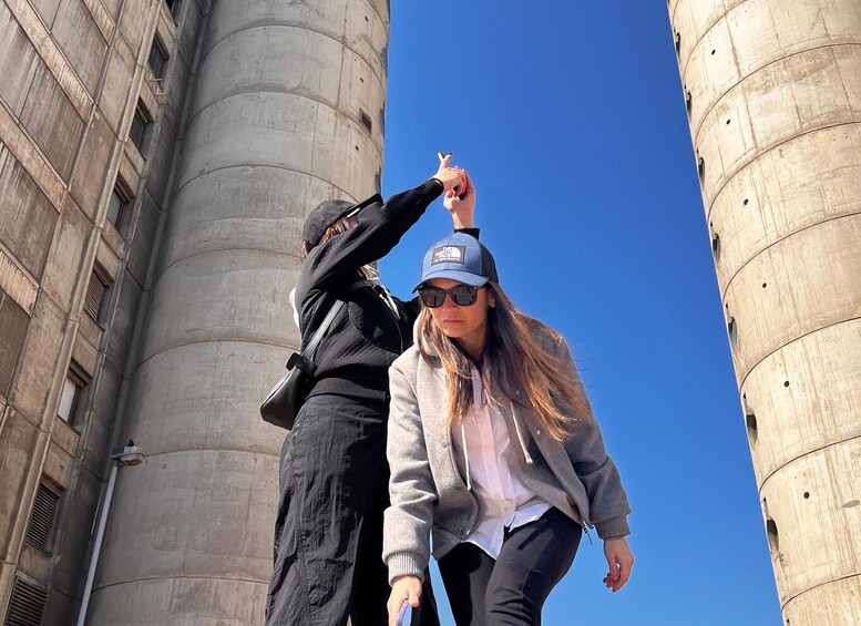 Two people standing in front of large cylindrical structures, possibly industrial or historical buildings. 1