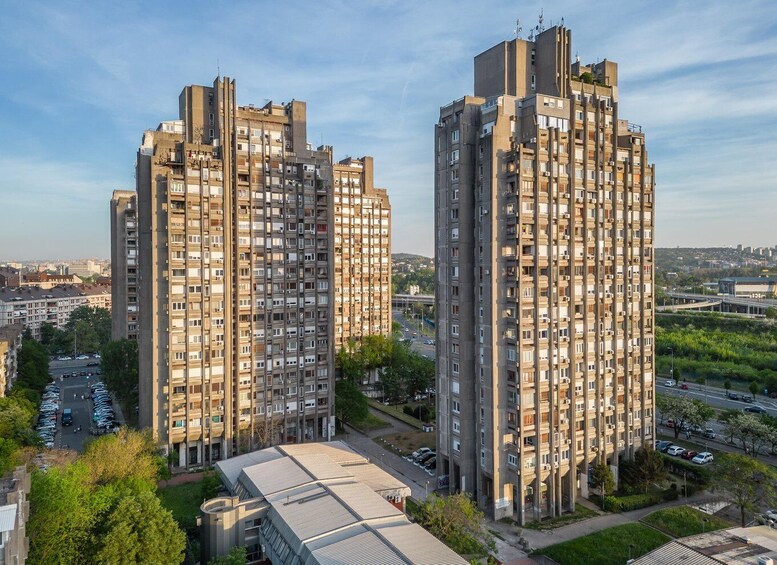 Two high-rise residential buildings with balconies, surrounded by greenery and a busy road. 3