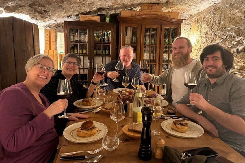 A group of people sitting around a wooden table in a rustic stone-walled room, toasting with wine glasses. 3