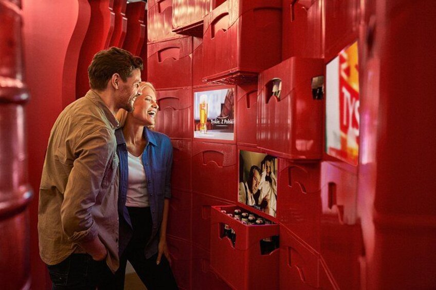 A couple standing in front of red lockers with photos and a TV screen. 4