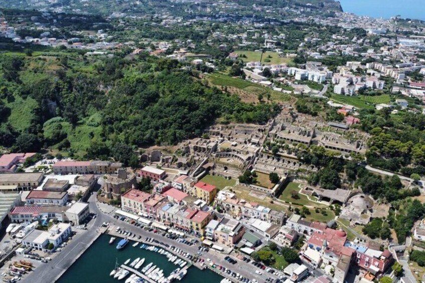Vista aérea de un pueblo costero con ruinas históricas, un puerto deportivo y vegetación circundante. 1