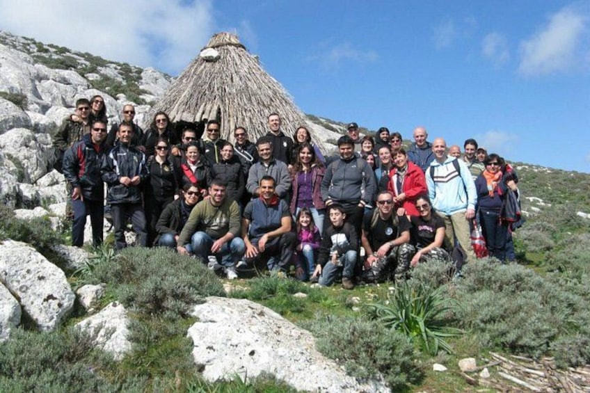 Un grupo de personas posando en un entorno rocoso al aire libre, con una estructura de techo de paja al fondo. 1