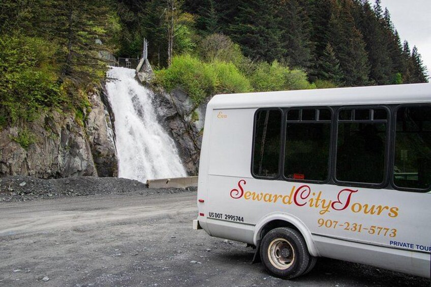 A bus with 'SewardCityTours' and a phone number parked near a waterfall. 2