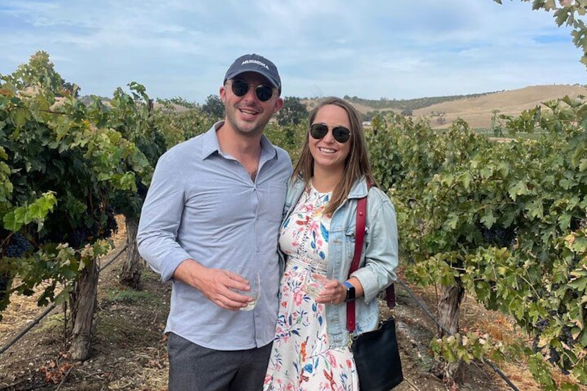 Two people standing in a vineyard with rows of grapevines. 3