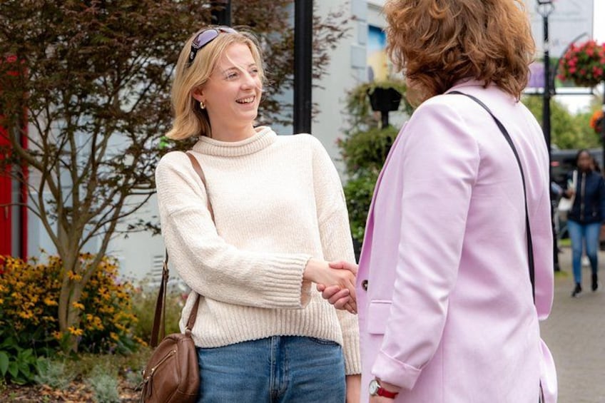 Two people holding hands, one in a white sweater and the other in a pink coat, standing on a sidewalk with trees and flowers in the background. 4
