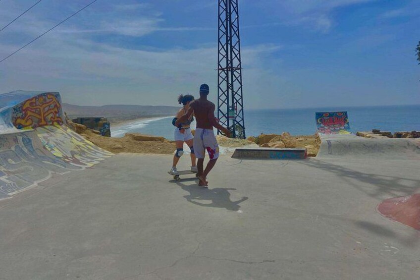 Een skatepark met hellingen en graffiti, een strand, en een hoge constructie op de achtergrond. 3