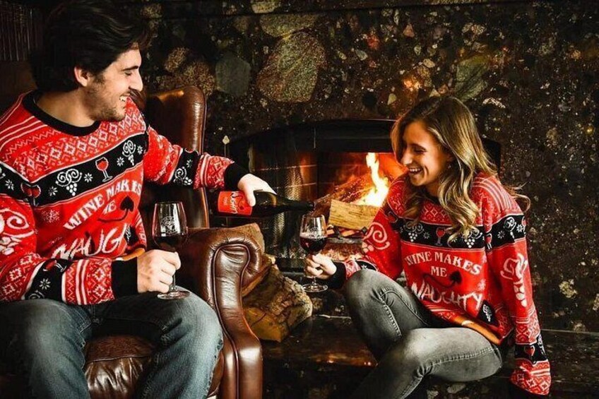 Two people in red sweaters with 'WINE MAKES ME' text, sitting by a fireplace. 1