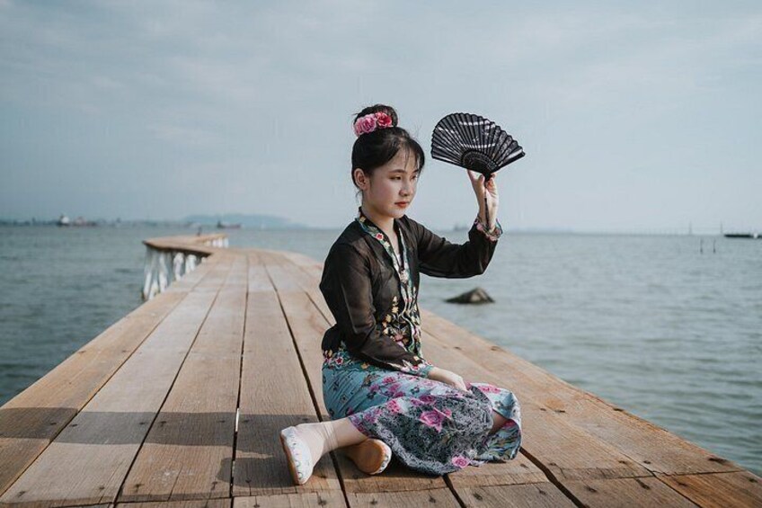 A person sitting on a wooden pier by the water, holding a fan. 5