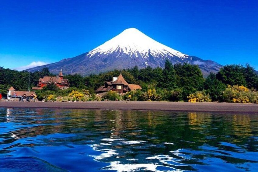 A snow-capped mountain, a body of water, and buildings along the shore. 1
