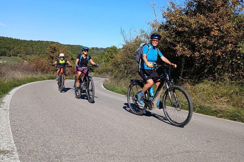 Three cyclists on a paved road with a hilly landscape in the background. 1