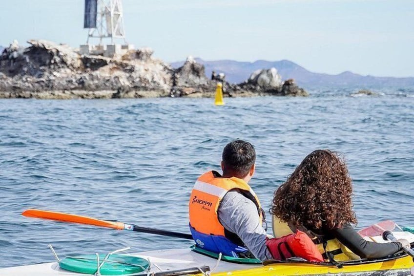 Two people in a kayak wearing life jackets, with a rocky coastline and a lighthouse in the background. 4