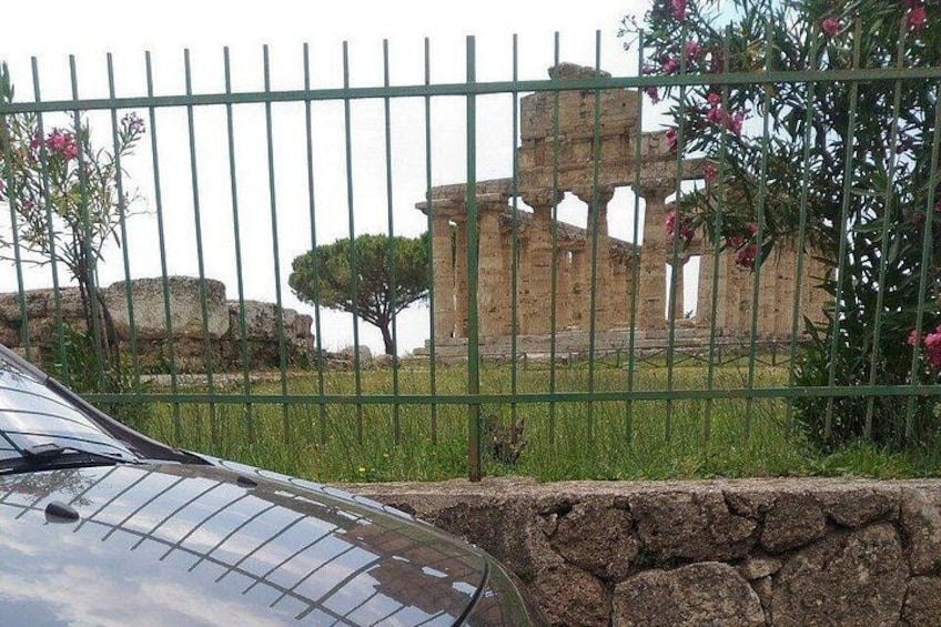 Une voiture est garée devant un mur de pierre, avec une clôture en métal et des ruines antiques en arrière-plan. 1