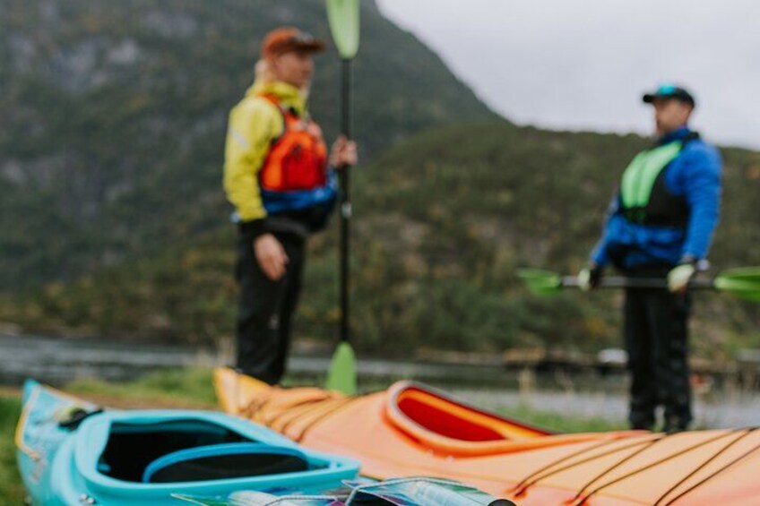 A person in a yellow jacket holding a paddle, a person in a blue jacket with a green life jacket, and a kayak in the foreground. 2