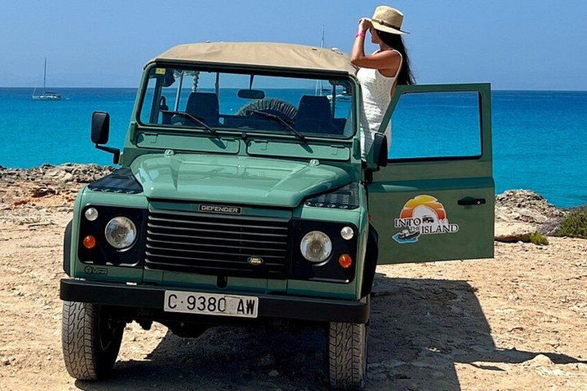 A green Defender vehicle parked on a rocky beach with a clear blue sea in the background. 4