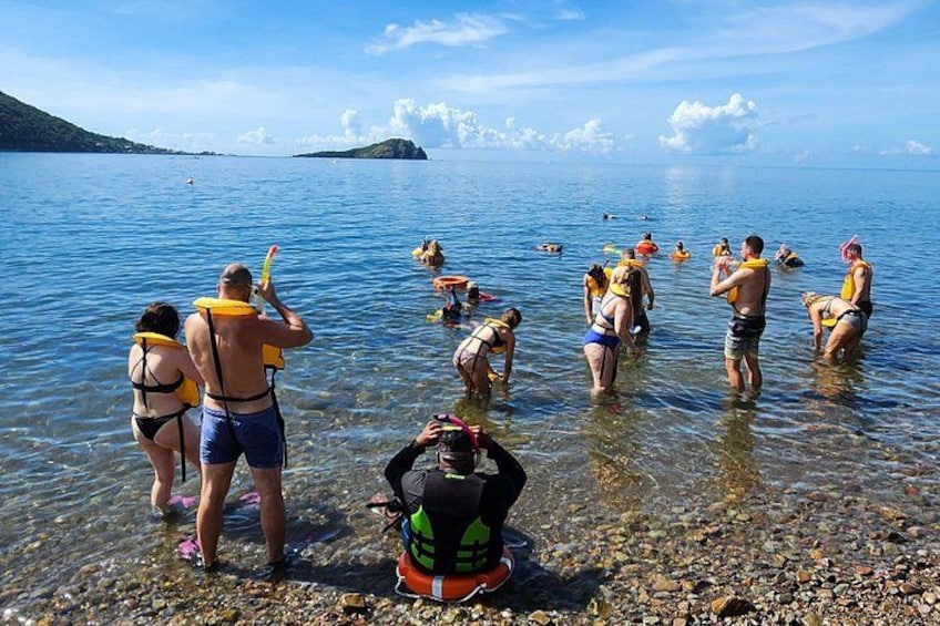 Personas de pie en aguas poco profundas, algunas con chalecos salvavidas, cerca de una costa rocosa con una vista despejada del agua y la tierra distante. 2