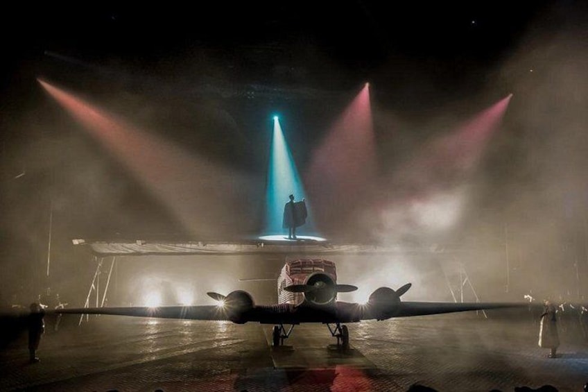 A stage with a person standing under a spotlight, surrounded by fog and a large aircraft. 3