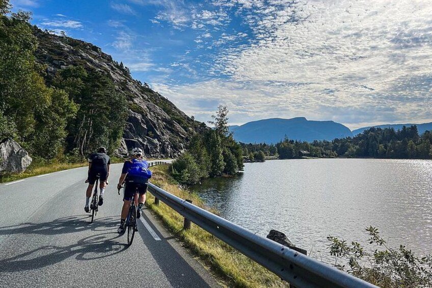 To cyklister på en asfalteret vej ved siden af en sø med bjerge i baggrunden. 4