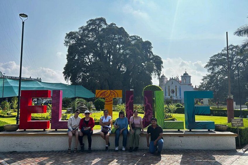 A group of people sitting on a bench in a park with colorful letter sculptures and a large tree in the background. 2