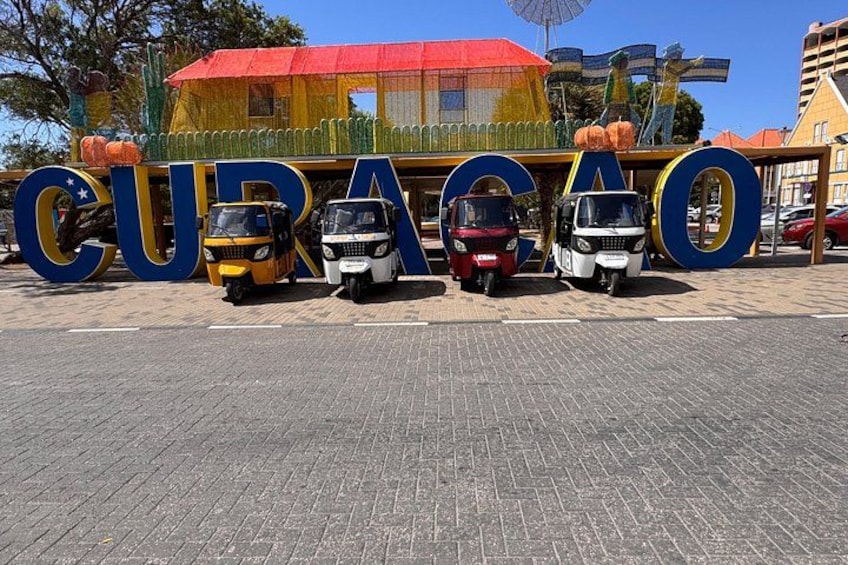 Un cartel con la palabra 'CURACAO' y tres tuk-tuks estacionados al frente. 3