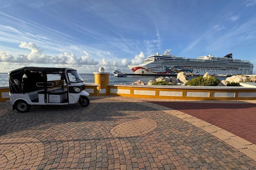 A tuk-tuk parked on a paved area with a cruise ship in the background. 2