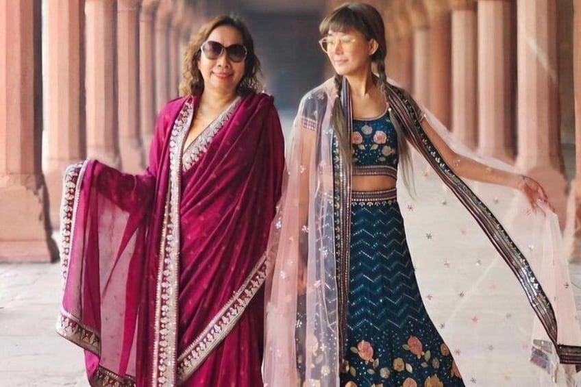 Due persone in abiti tradizionali, una con un sari color borgogna e l'altra con un vestito blu, si trovano in un contesto architettonico con colonne. 3