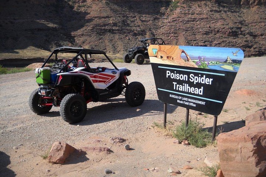 A desert trailhead sign with a vehicle parked nearby. 4