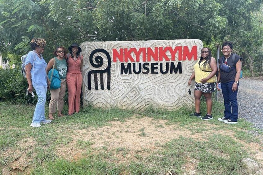 Un groupe de personnes se tient devant une enseigne indiquant l’entrée du musée Nkynkym. 3