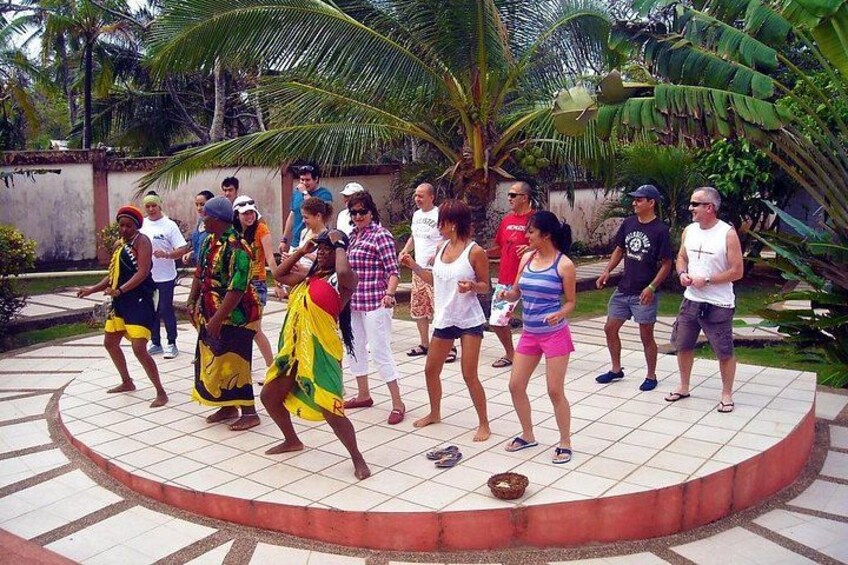 A group of people dancing in a circular outdoor area with tiled flooring and palm trees in the background. 1