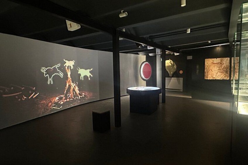 A museum interior with a projection of prehistoric animals, a circular display, and a glass case with artifacts. 2