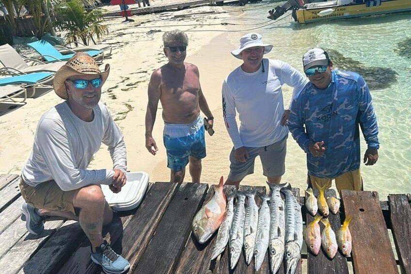 Four people standing on a wooden dock with a wooden table displaying various fish. 4