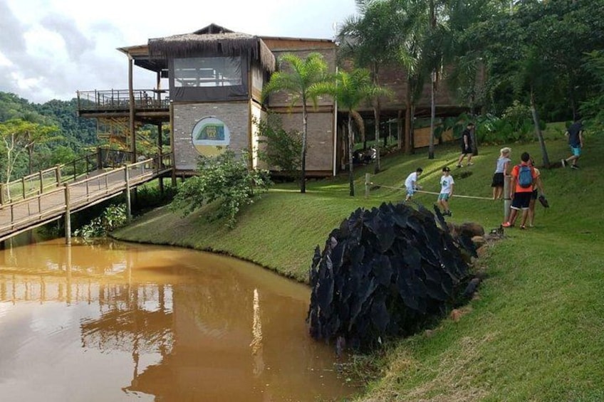 A building with a viewing platform, a pond, and people walking on a grassy area. 1