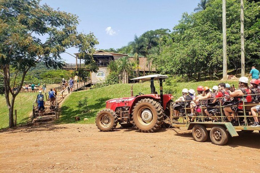 A red tractor pulling a cart with people on it, a dirt path, and a building in the background. 2