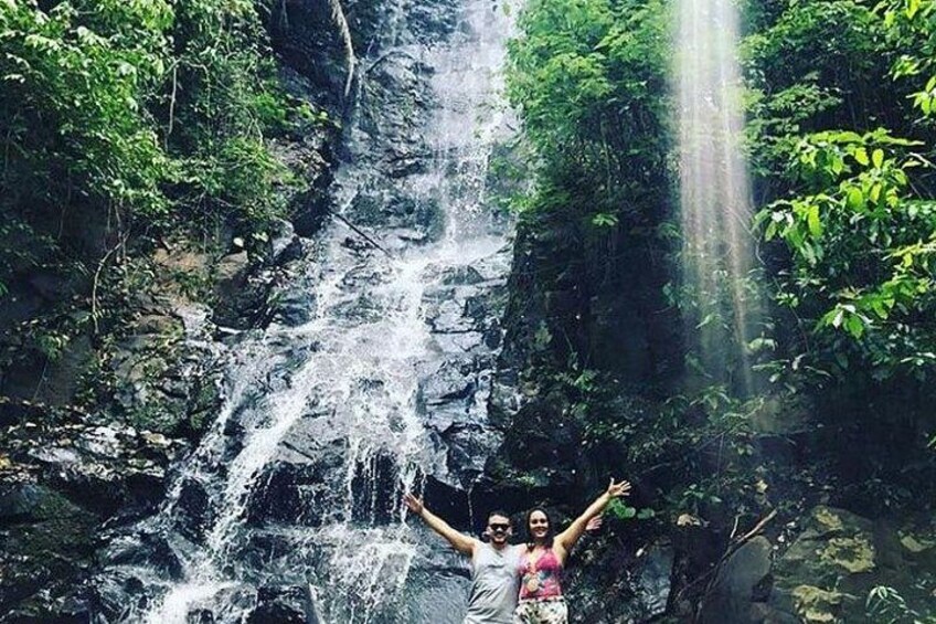 A waterfall in a forested area with lush greenery and a person standing with arms raised. 1
