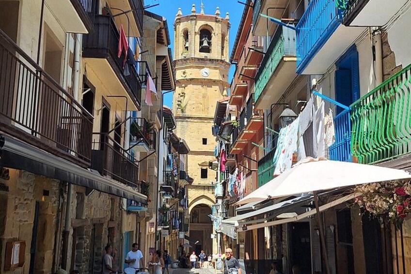 A narrow street with buildings, a bell tower, and people walking. 3