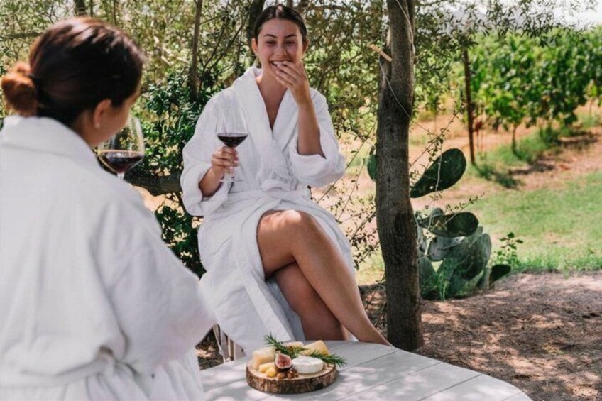 Two people in white robes sitting at a table with a basket of food and a glass of red wine in a wooded area. 3