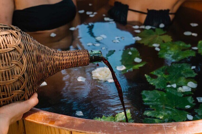 A person pouring a dark liquid from a wicker bottle into a copper basin filled with water and lily pads. 1