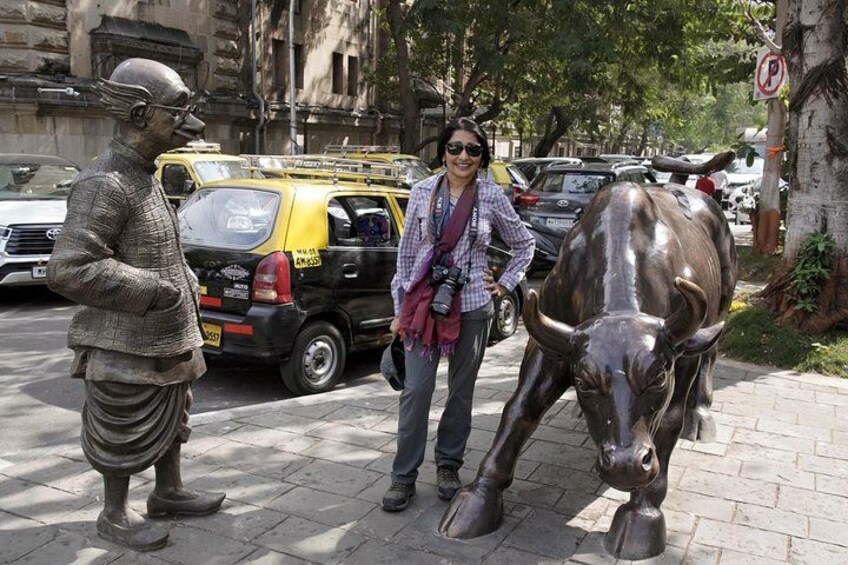 A person standing next to a bull statue on a city street with cars and buildings in the background. 5