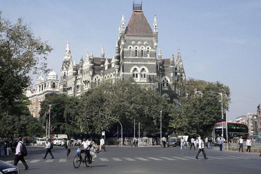 A busy street with pedestrians, cyclists, and a red bus. In the background, there is a large, ornate building with multiple spires and a clock tower. 3