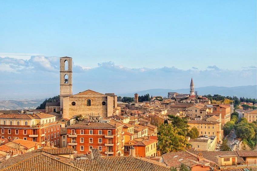 A historic town with a prominent bell tower, a church with a tall spire, and a mountainous backdrop. 1
