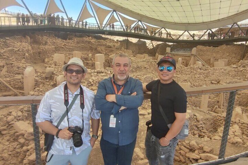 Three people standing in an archaeological site with exposed ruins and a large tent structure in the background. 3
