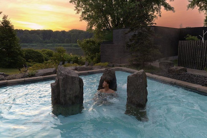 Une personne dans un bain à remous, avec des rochers dans l’eau, entourée d’un jardin aménagé et offrant une vue sur des arbres et un coucher de soleil. 1