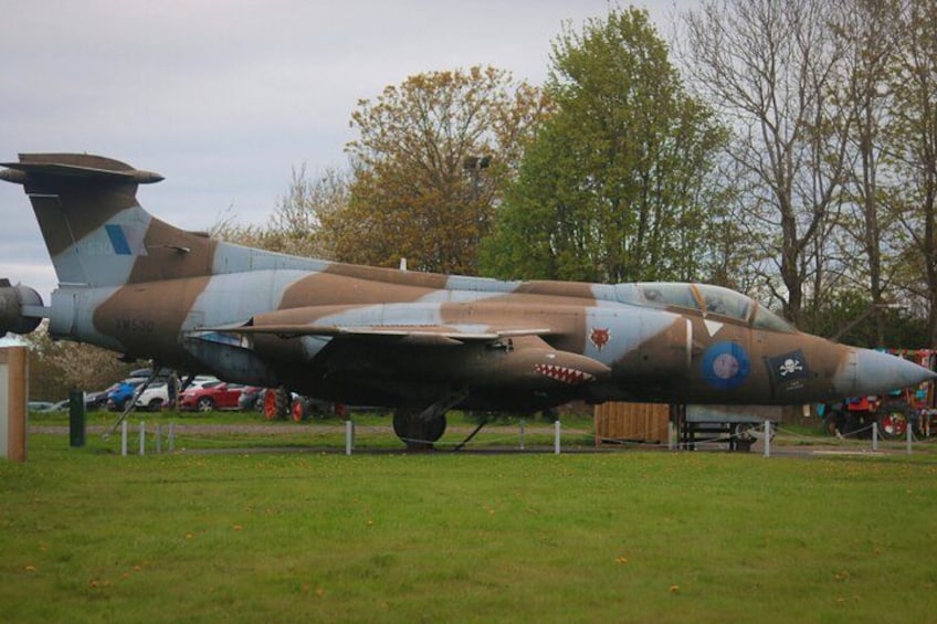 Un avion militaire est exposé dans un champ herbeux, avec des arbres et un bâtiment en arrière-plan. 3