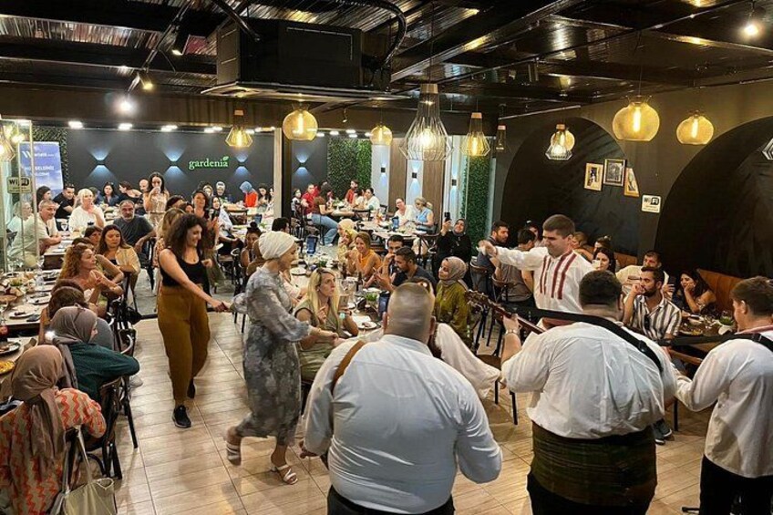 Un comedor interior concurrido, personas sentadas en mesas, alguien bailando y un letrero que dice 'gardenia'. 1