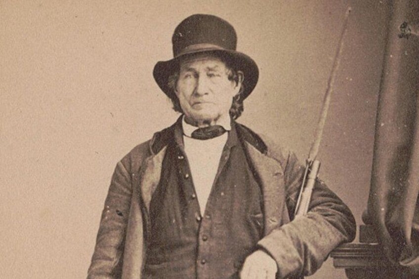 A person in historical attire holding a rifle, standing in front of a flagpole. 1