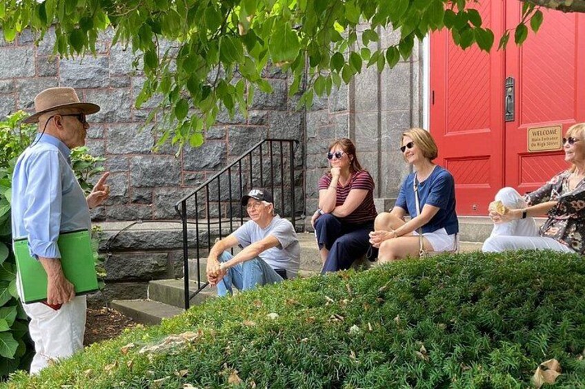 A group of people sitting on steps in front of a red door, with greenery and stone walls in the background. 5
