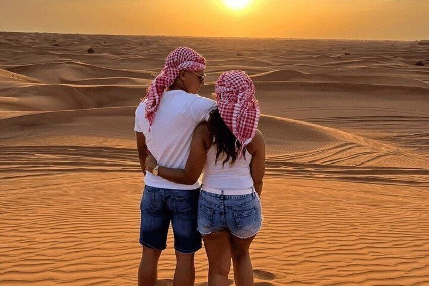 A couple standing in a desert with sand dunes and a sunset in the background. 5